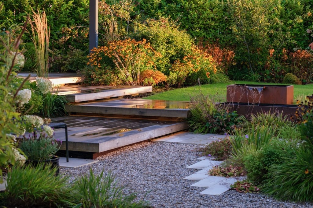 Large deck steps lead from a path and gravel area to seating deck. Surrounded in planting with a water feature nearby